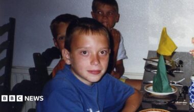 Anton wearing a blue t shirt on the left. There are several other people smiling at the camera as they sit down at a dinner table. There are green and yellow napkins on plates.