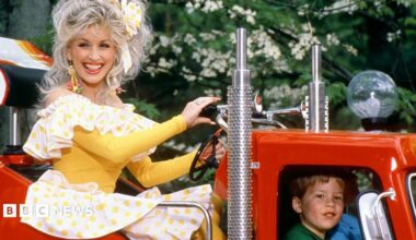 Dolly Parton is pictured riding on a truck at her famous Dollywood theme park in Tennessee in 1977, she's in a yellow and white dresss