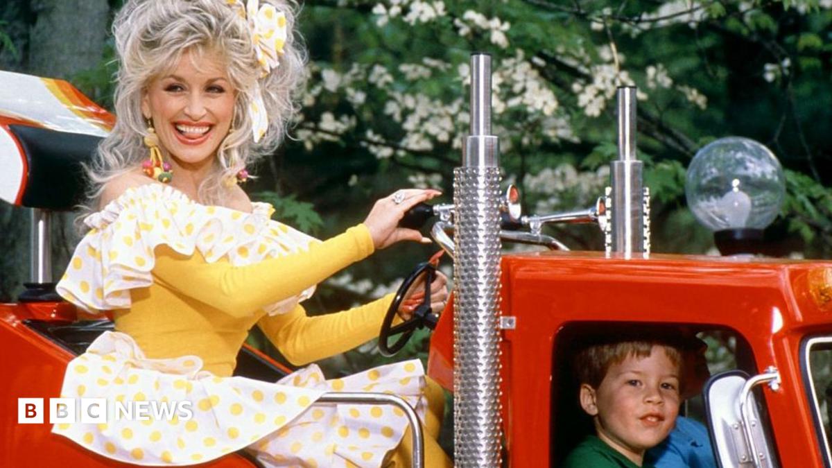 Dolly Parton is pictured riding on a truck at her famous Dollywood theme park in Tennessee in 1977, she's in a yellow and white dresss