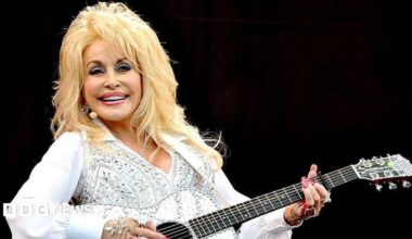 doll parton in a white rhinestone top and a matching acoustic guitar. she has blonde hair and full make up and a big smile as she stands on stage in front a dark background
