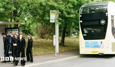 A yellow bus passing a bus stop as a group of men in black outfits stand by a pus stop.