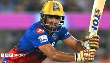 Mahipal Lomror in a blue and red shirt, with gold gloves and helmet, hits a shot with a bat facing upwards