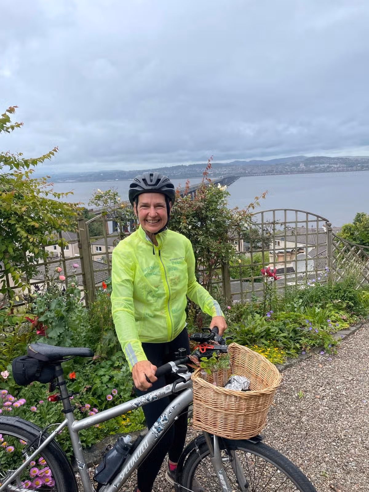 An image of a woman wearing a helmet and neon yellow jacket, standing in a garden next to a bike.