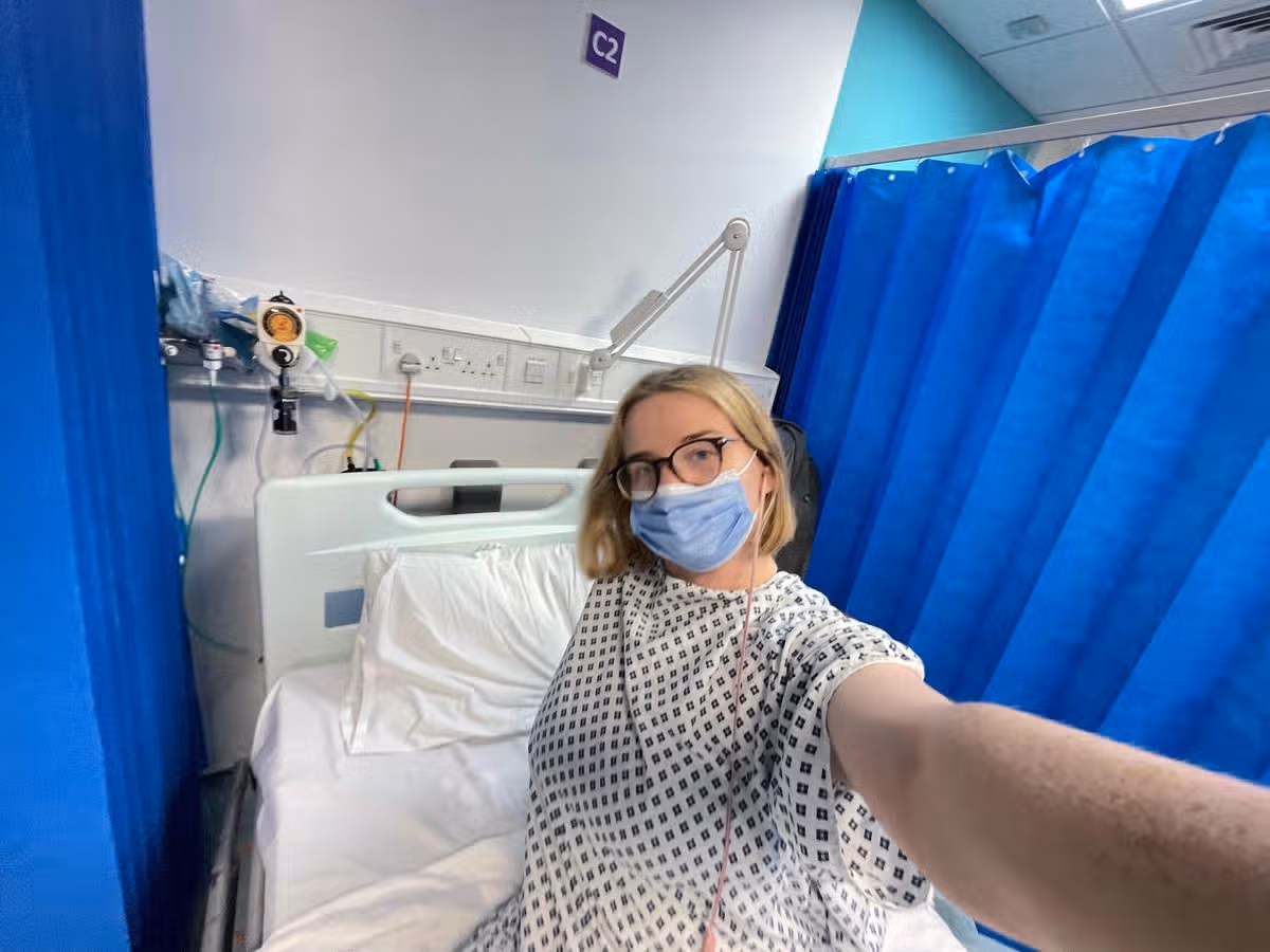 A young woman in hospital taking a selfie while wearing a surgical gown and face mask