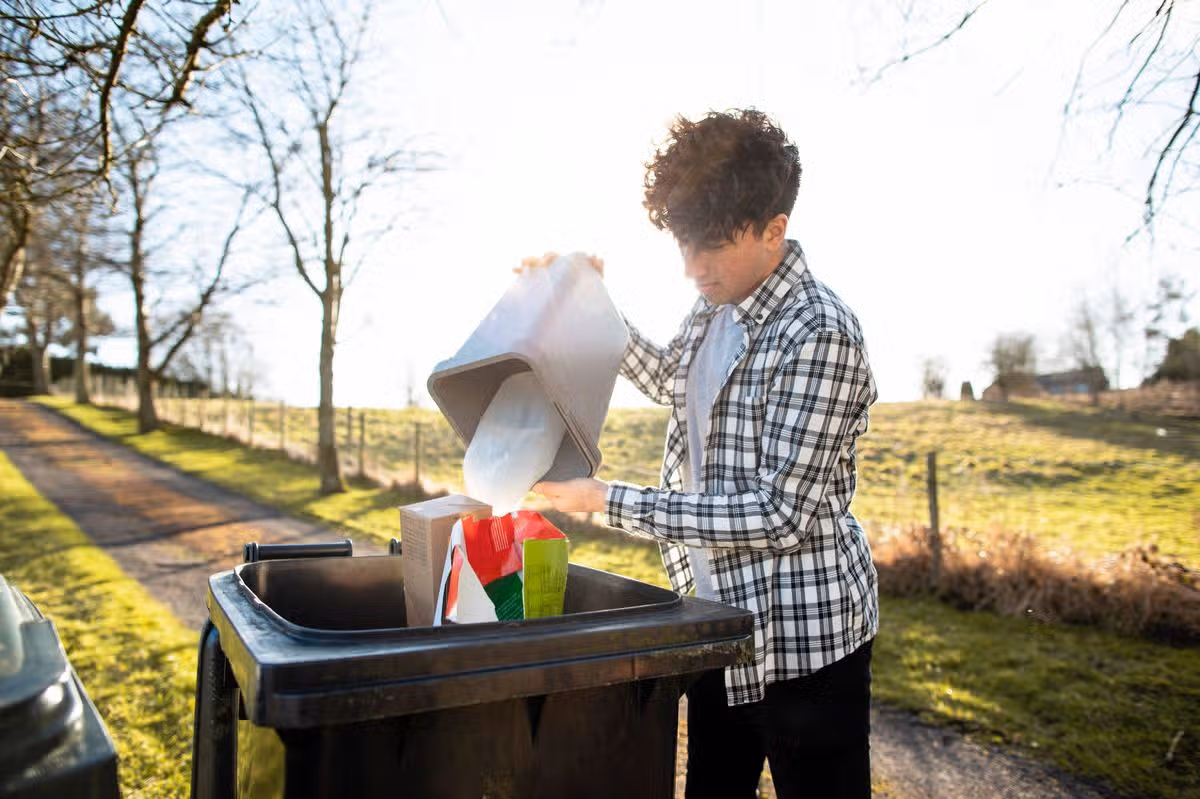 A teenage boy pouring recycling waste into the bin out of bag at the bottom of his drive. He is in the North East of England.