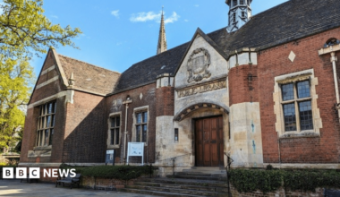An old stone and brick building, which says Public Library at the front, with a large number of black, small paned windows. There are steps up to the entrance, with a large wooden door. There are two signs and a green tree to the left. Bushes are out the front.