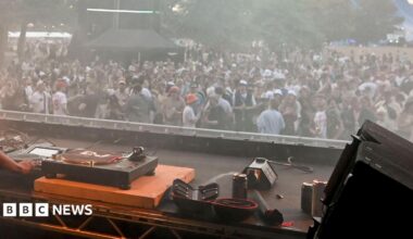 A set of DJ decks in the foreground with a crowd of people in the background, at a music festival. There's trees in the background behind the crowd. The crowd themselves are dancing, all are wearing various items of summer gear like shorts, T-shirts and bucket hats.