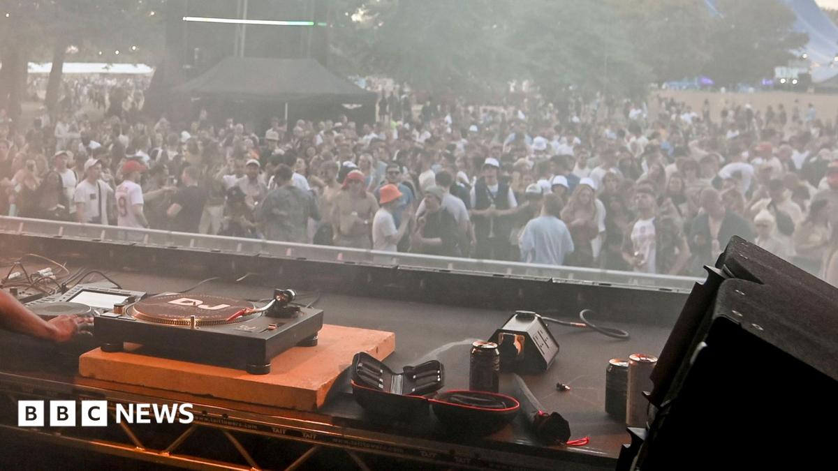 A set of DJ decks in the foreground with a crowd of people in the background, at a music festival. There's trees in the background behind the crowd. The crowd themselves are dancing, all are wearing various items of summer gear like shorts, T-shirts and bucket hats.