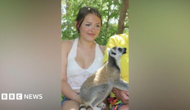 Lillie Nichols has tied back brown hair, a white vest, denim shorts with a ring-tailed Lemur on her knee.