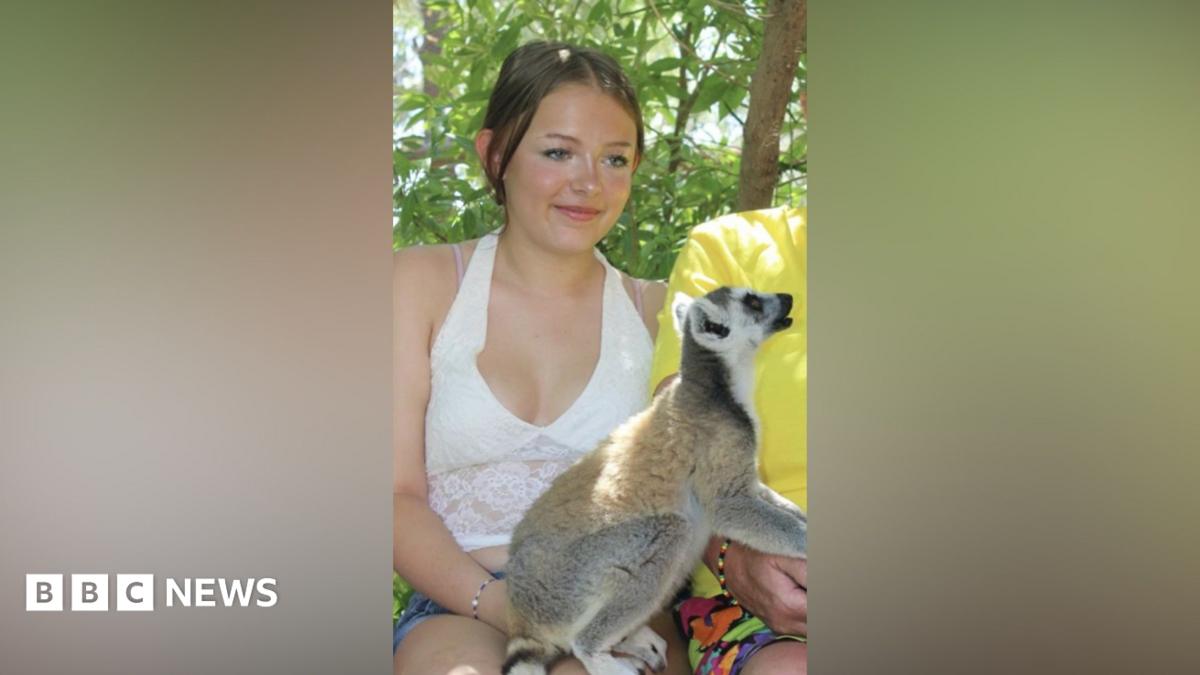 Lillie Nichols has tied back brown hair, a white vest, denim shorts with a ring-tailed Lemur on her knee.