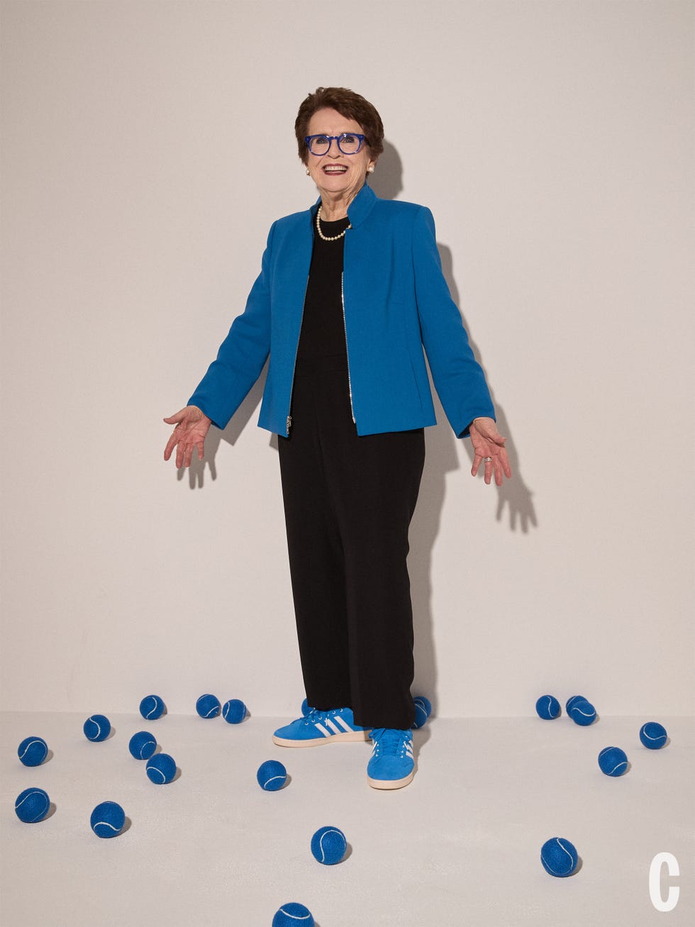 Billie Jean King in a blue blazer and matching sneakers stands among scattered blue tennis balls, smiling with arms outstretched. Billie Jean King in a blue blazer and matching sneakers stands among scattered blue tennis balls, smiling with arms outstretched.