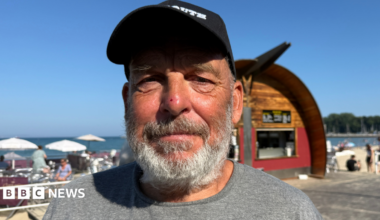 A man wearing a cap and a grey t-shirt looking into the camera on a sunny day next to the beach