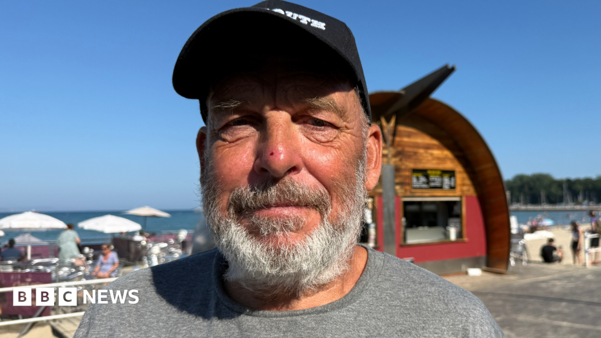 A man wearing a cap and a grey t-shirt looking into the camera on a sunny day next to the beach