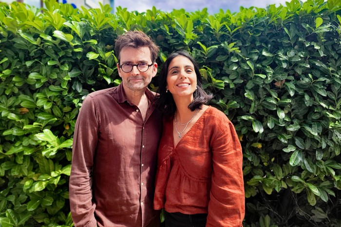 David Tennant and Anjli Mohindra smiling at the camera, in front of a leafy background