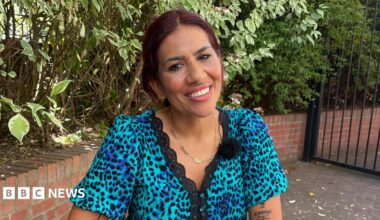 A woman with dark hair and a blue animal print dress smiles sitting in a garden.