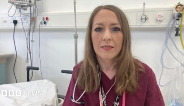A woman wearing maroon hospital scrubs. She is in a hospital ward and is looking at the camera. She has long brown hair.
