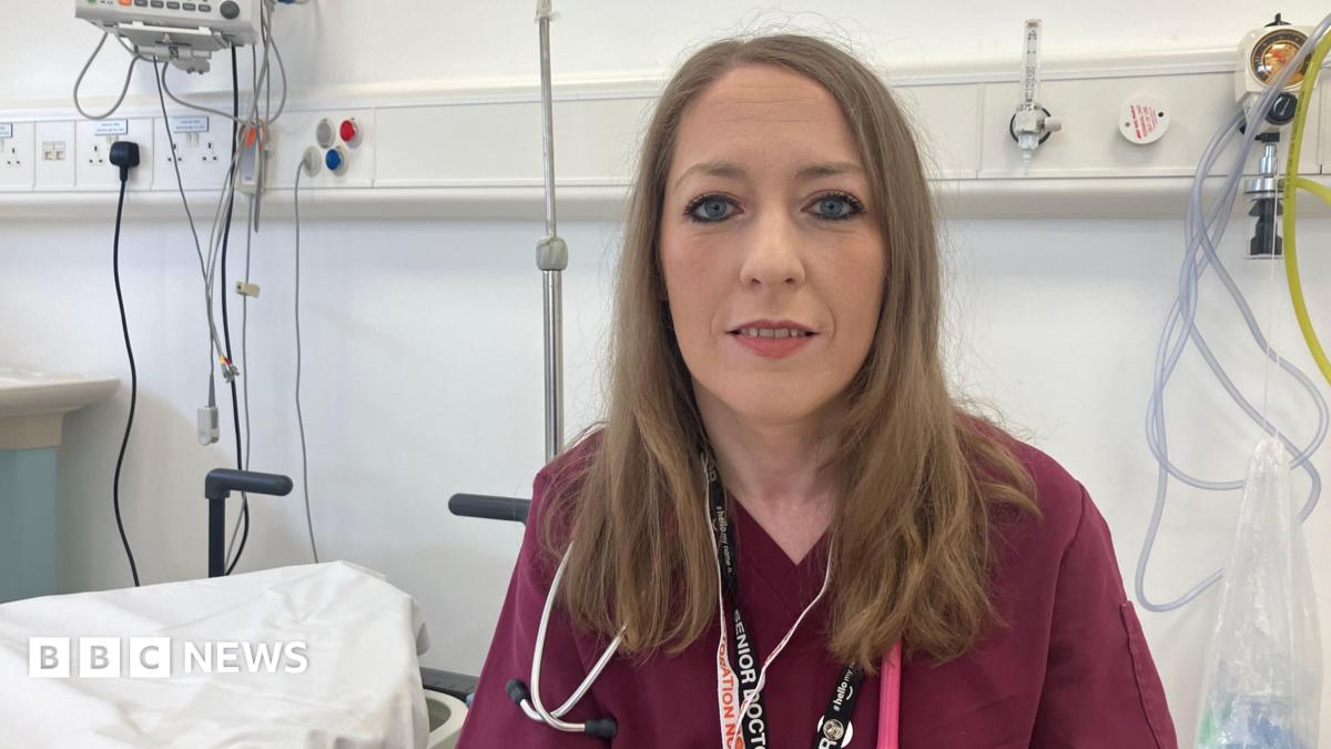 A woman wearing maroon hospital scrubs. She is in a hospital ward and is looking at the camera. She has long brown hair.
