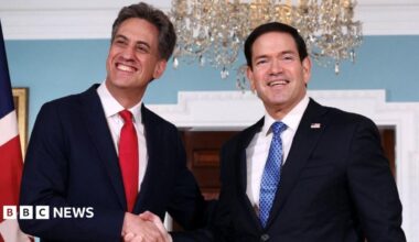 Two men with dark hair shake hands while smiling. They are wearing business suits