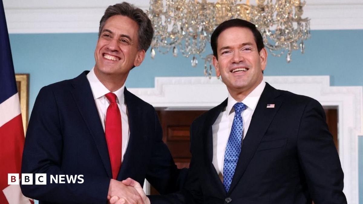 Two men with dark hair shake hands while smiling. They are wearing business suits