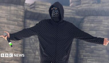 EsDeeKid performs in front of a blow-up castle during a set in Illinois.  He is seen looking at the crowds while holding a microphone and wearing a black mask with holes and a hoodie.