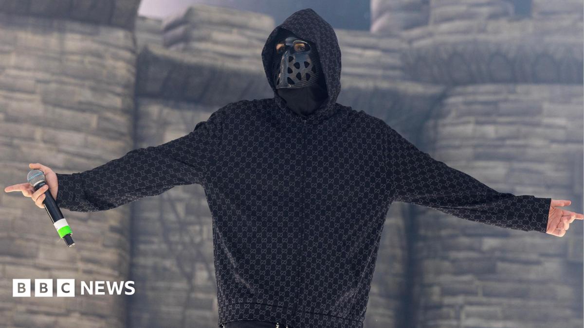 EsDeeKid performs in front of a blow-up castle during a set in Illinois.  He is seen looking at the crowds while holding a microphone and wearing a black mask with holes and a hoodie.
