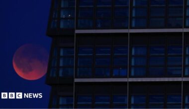 A red Moon lurking behind a glass skyscraper.