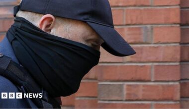 A close-up of the head and shoulders of a man with his face obscured by a black scarf and black cap as he walks towards a red-brick court. His eyes can be seen and his is looking downwards. He is wearing a blue suit jacket and the strap of a bag over his shoulder.