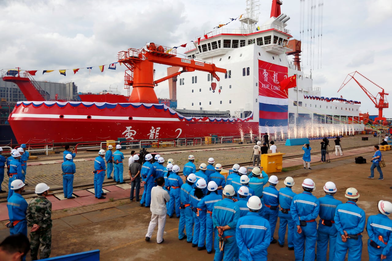 Many people in blue overalls and white helmets stand on a dock in front  of a large red and white ship.