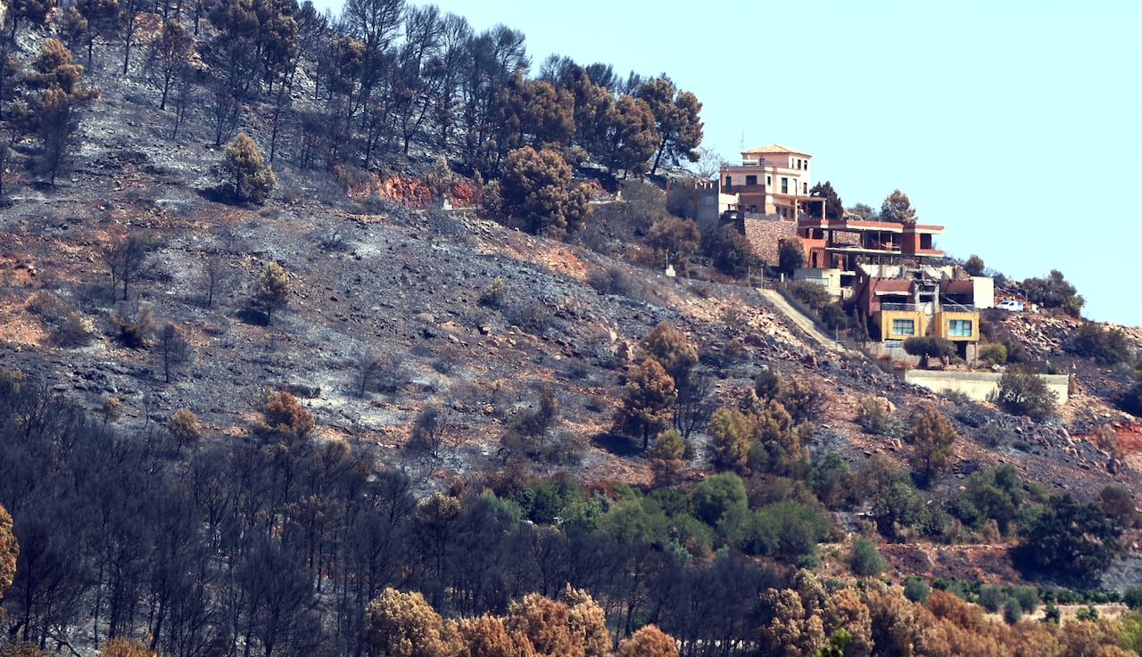 A cluster of houses sit next to a large area on a hill that has been scorched with fire, leaving it smoking and bare of foliage.