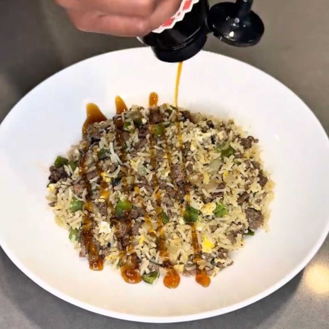 This photo shows an example of boy kibble: A white bowl is filled with rice, ground beef, green peppers and onion. A hand above the bowl is squeezing a bottle of brown-colored sauce into the bowl.