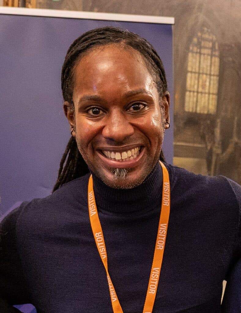 Man with braided hair wearing a navy turtleneck and an orange visitor lanyard, smiling at an indoor event
