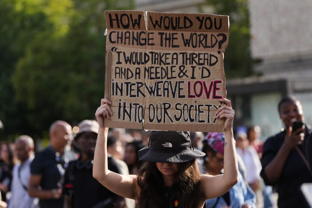 A protester holds a cardboard sign reading: How would you change the world? "I would take a thread and a needle & I'd interweave love into our society