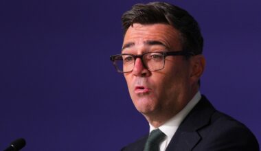 Andy Burnham, Mayor of Greater Manchester, speaking at a podium against a dark blue background.