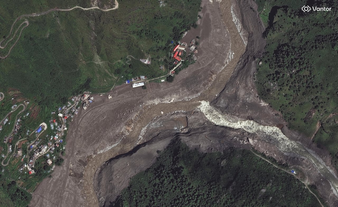 A satellite image shows a brown mud filled river flooding a village, nestled in a green valley.