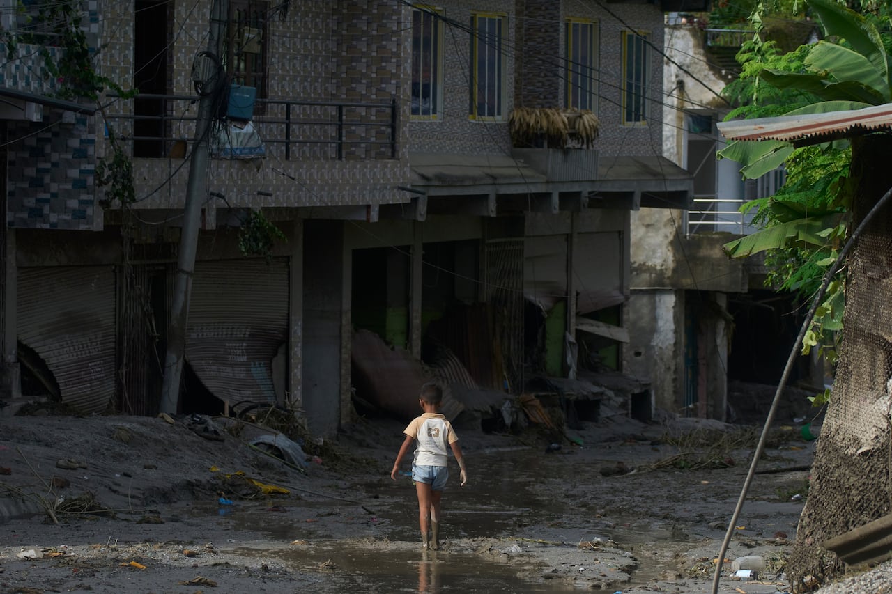 A child, with his back to the camera, walks down a mud and debris-covered lane between homes that are also covered in mud.
