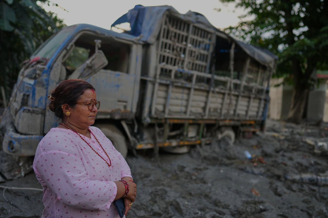 A woman in a pink top, with a red beaded necklace around her neck and matching bracelet, stands near a truck that has been covered in mud from a flash flood.