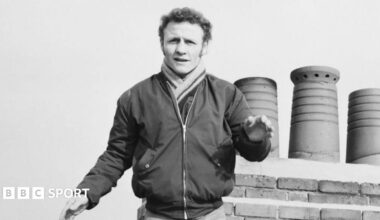 Paddy Maguire holds up his arm and stands in front of a row of chimneys in 1973