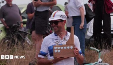 Alexandre Voisine in cycling clothing and a cap, holding a wooden trophy depicting a penny farthing and smiling.