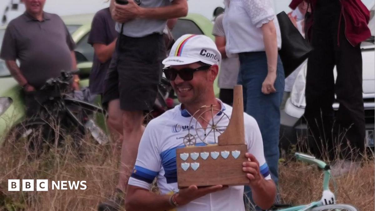Alexandre Voisine in cycling clothing and a cap, holding a wooden trophy depicting a penny farthing and smiling.