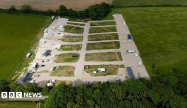 A view of the field. About six caravans are parked on hardstanding along with other vehicles and yellow diggers. There is a pylon in the middle of the field.