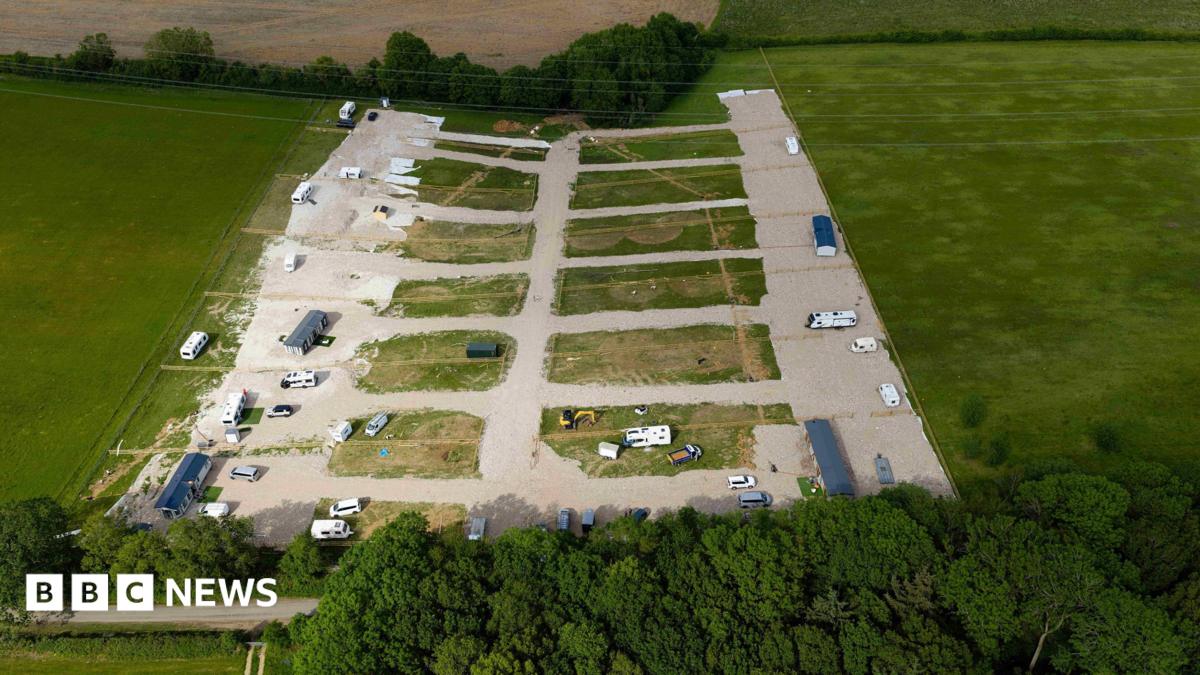 A view of the field. About six caravans are parked on hardstanding along with other vehicles and yellow diggers. There is a pylon in the middle of the field.