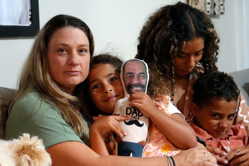 A woman sits on a couch with her three kids. One of the girls holds a doll with her father's likeness printed on it.