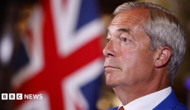Nigel Farage is wearing a blue suit, white shirt and a tie. He has short grey hair. A Union flag can be seen in the background.