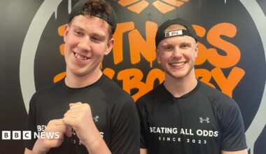 Shay and Bobby are both wearing black T-shirts and caps facing backwards. They are standing in front of a black and orange Fitness By Bobby logo.