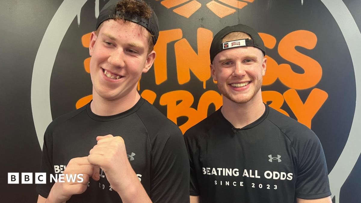 Shay and Bobby are both wearing black T-shirts and caps facing backwards. They are standing in front of a black and orange Fitness By Bobby logo.