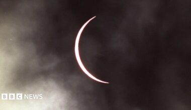 The moon has moved across the sun leaving a thin band of the sun visible which has taken on a pink hue. The sky is cloudy.