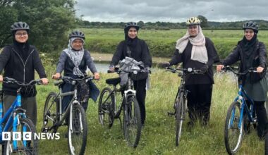 Five women dressed in dark clothing and wearing hijabs and bicycle helmets stand holding five bikes. They are on a grass verge alongside a river, with a grassy field in the background, lined by trees
