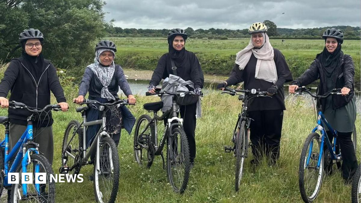 Five women dressed in dark clothing and wearing hijabs and bicycle helmets stand holding five bikes. They are on a grass verge alongside a river, with a grassy field in the background, lined by trees