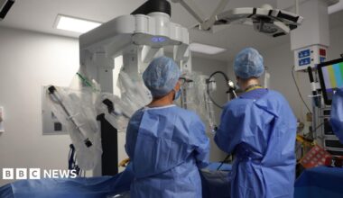 A woman wearing a light-coloured checked jacket and an NHS lanyard, standing in a hospital room. Medical equipment, a curtained area and information boards are in the background.