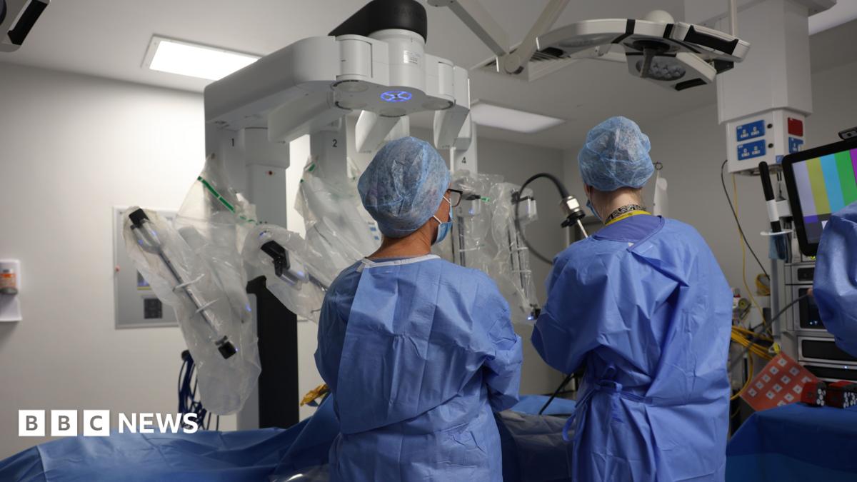 A woman wearing a light-coloured checked jacket and an NHS lanyard, standing in a hospital room. Medical equipment, a curtained area and information boards are in the background.